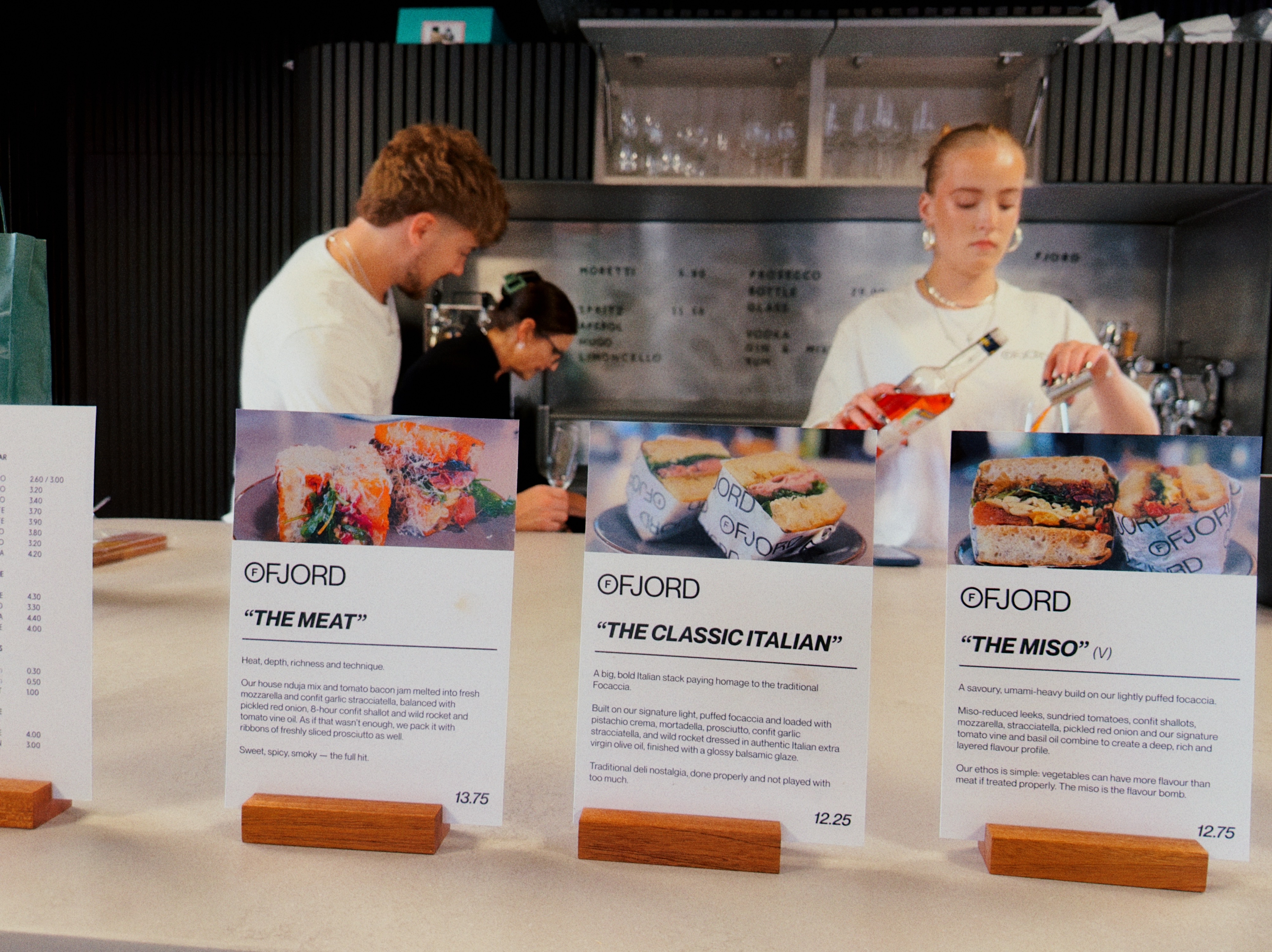 Two Fjord team members working behind the counter, one pouring a spritz, with sandwich menu cards along the front.